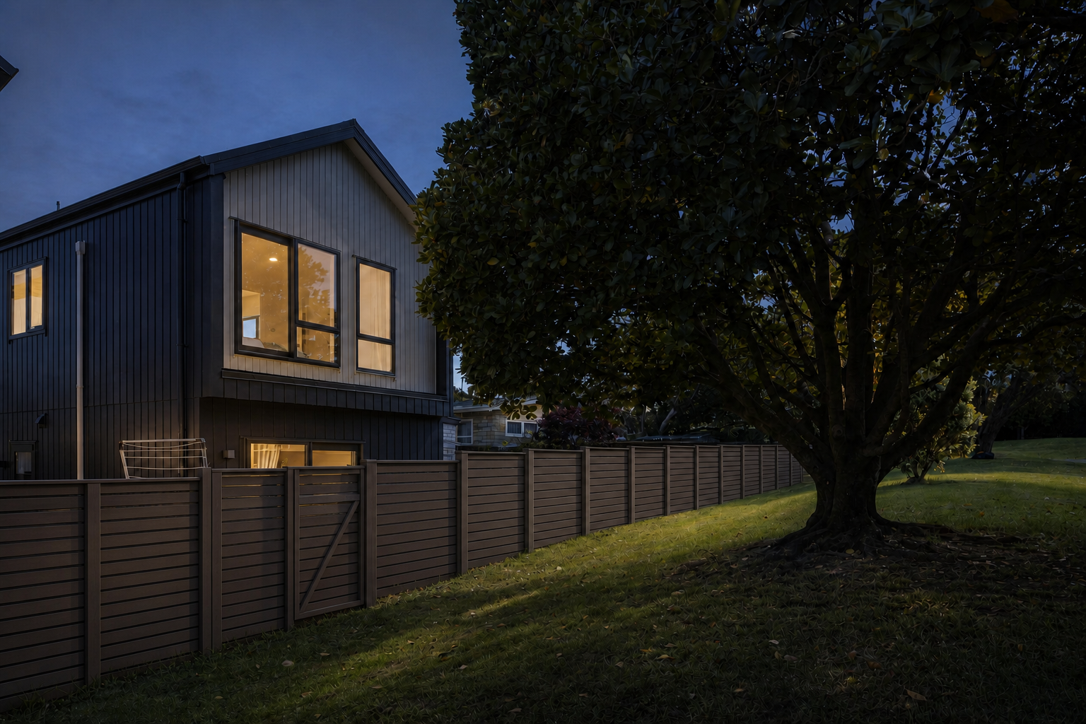 Garden-side evening view of Stonefield Residence with mature tree and dark timber fence.